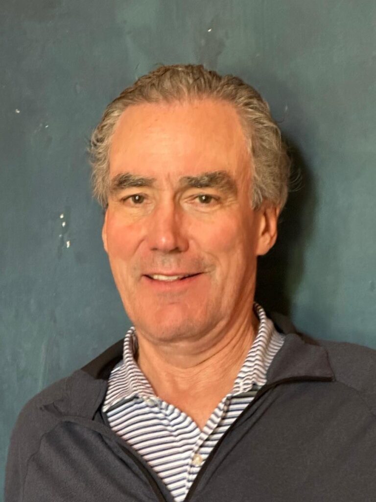 Dr. David Systrom with gray, wavy hair is standing in front of a solid dark teal wall. He is smiling slightly and wearing a navy blue zip-up jacket over a light blue and white striped collared shirt.