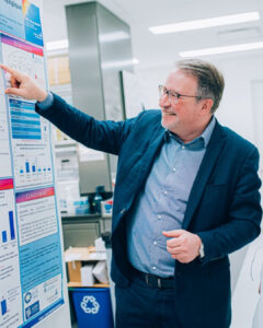 Dr. Alain Moreau, wearing a blue suit and glasses, smiles as he points to a scientific poster in a research lab.